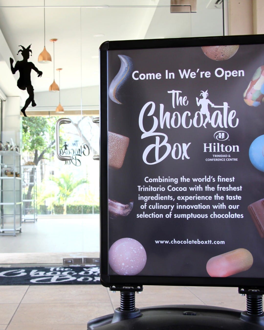 A sign for The Chocolate Box at Hilton Trinidad advertises their open status and highlights handcrafted chocolates made with Caribbean cocoa. The shop interior is visible in the background. | Trinitario